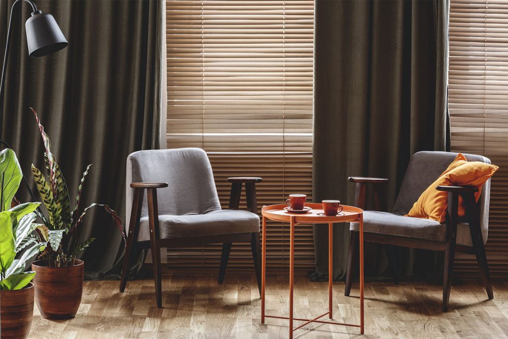 Vintage armchairs, orange coffee table with two cups, plants standing by the window with curtains and blinds in a living room interior